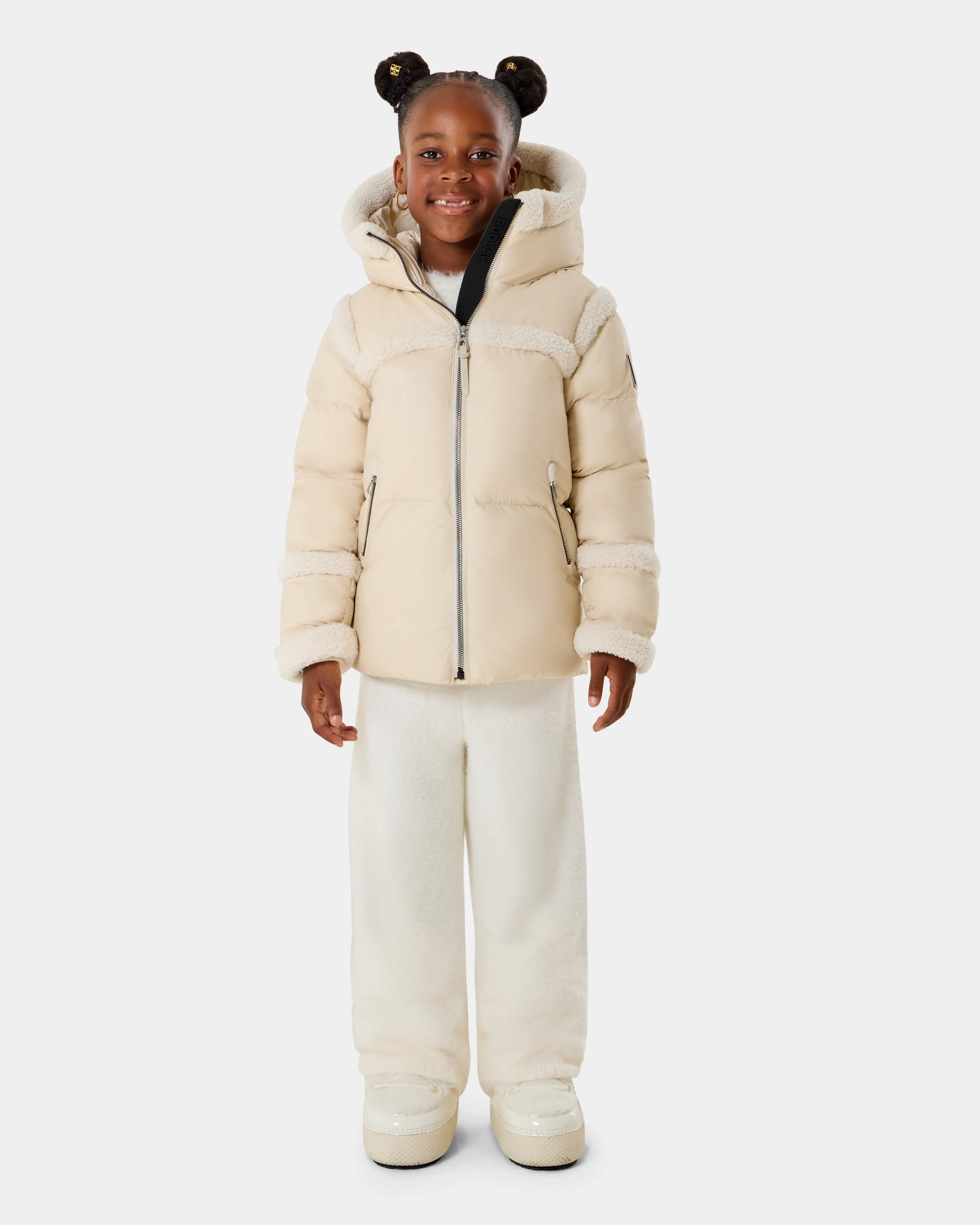 A smiling young girl with two buns stands facing forward against a white background, wearing the Rudsak JULIANA K - K125550 Vanilla light beige hooded puffer jacket, paired with white pants and boots.