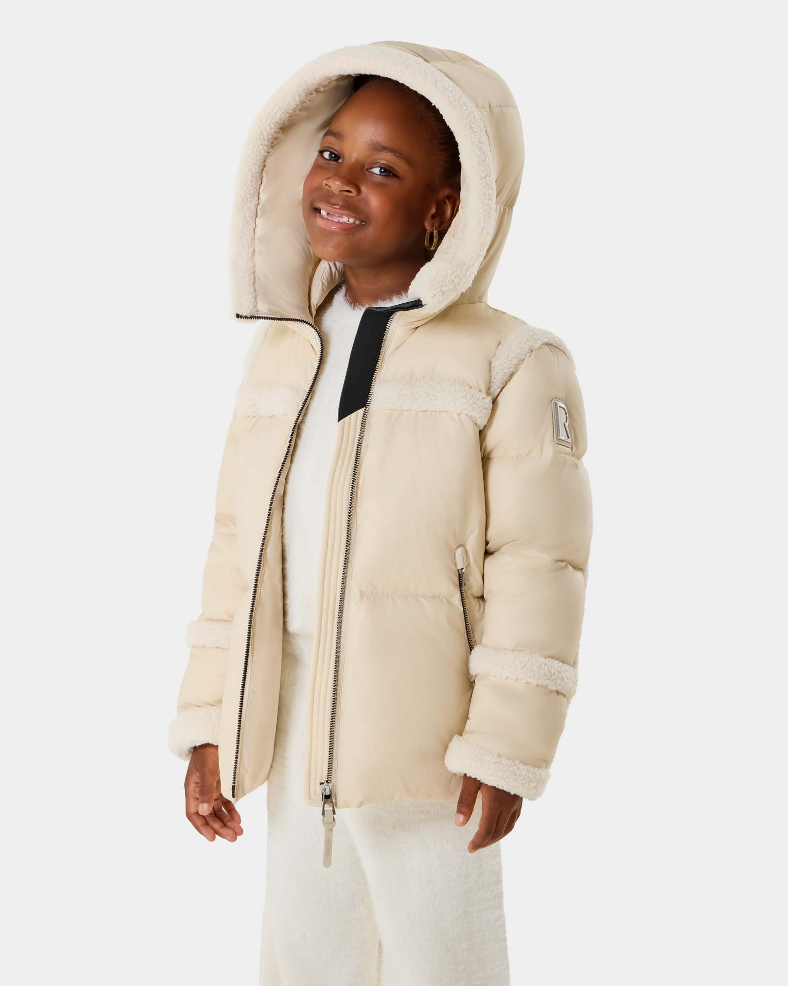 A young girl smiles in the Rudsak JULIANA K - K125550 Vanilla puffer jacket with a hood and fuzzy trim, matched with cream pants and sweater, standing against a plain white background.