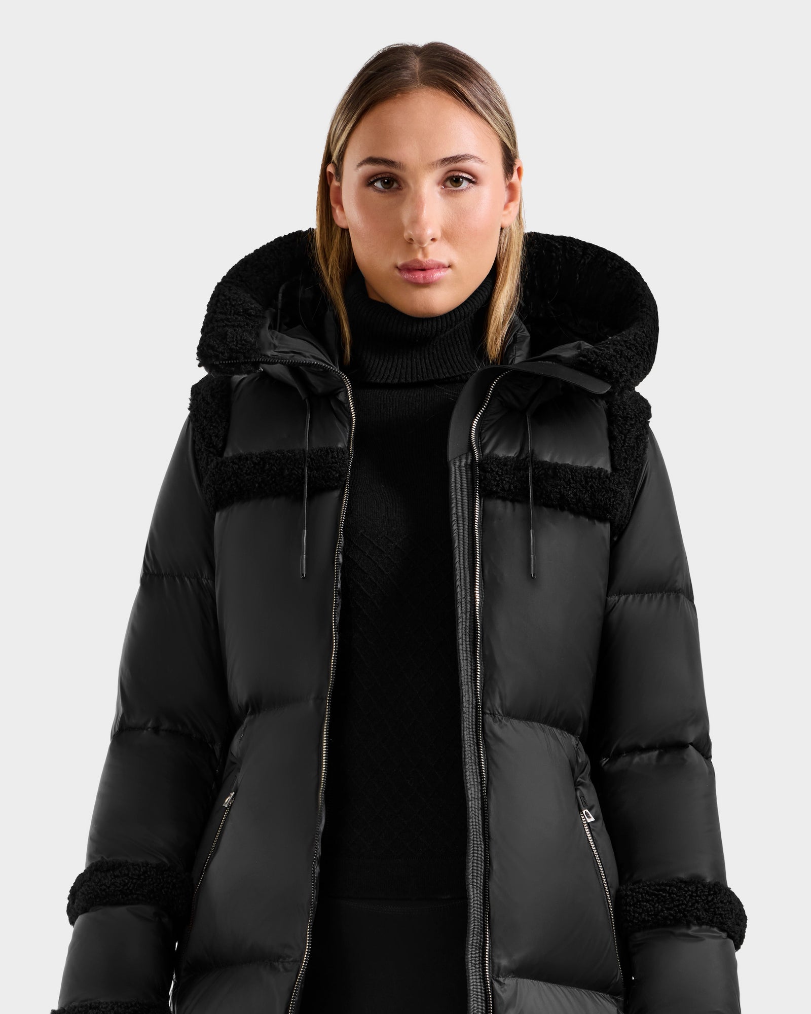 A woman wears the Rudsak JULIANA L - L125535 Black/Black down puffer jacket with hood and a black turtleneck, standing against a plain light background—showcasing chic style and warmth.