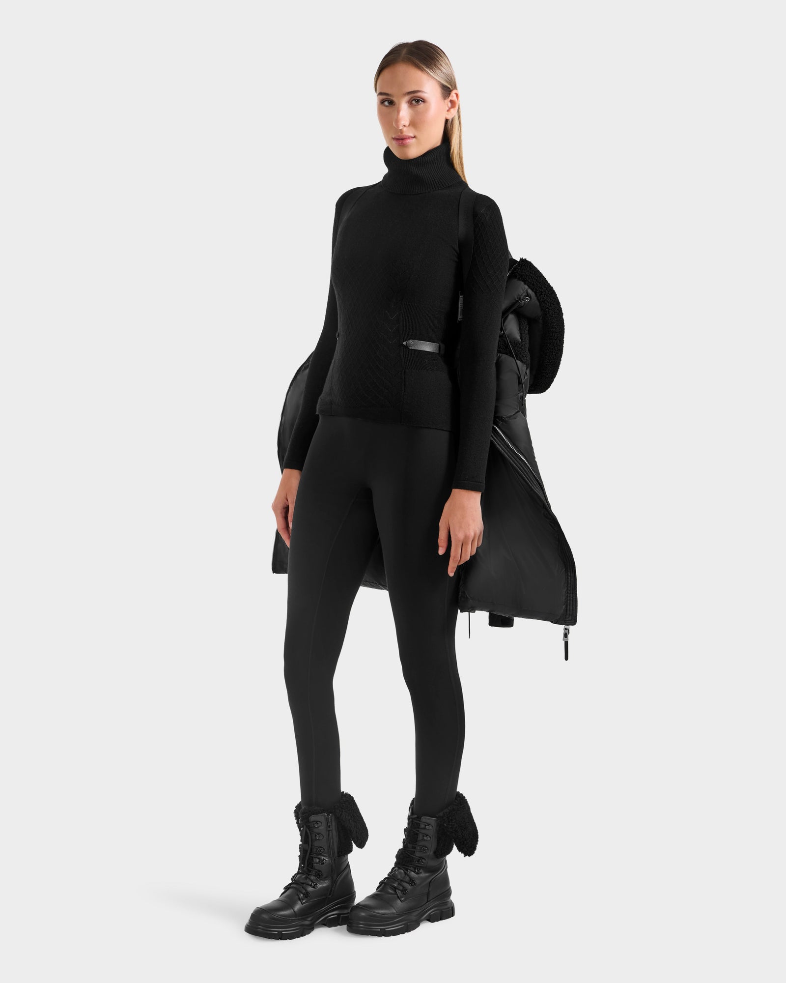 A woman stands facing forward against a plain, light background, wearing the Rudsak JULIANA L - L125535 Black/Black down puffer draped over her shoulders, paired with a black turtleneck sweater, leggings, and lace-up boots.