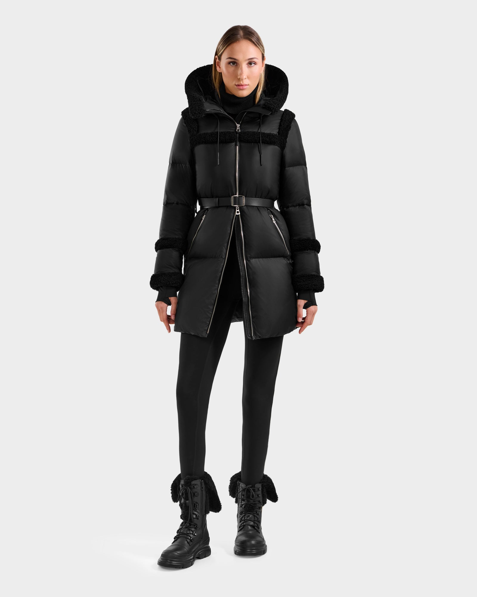 A woman faces forward in a Rudsak JULIANA L - L125535 Black/Black belted puffer coat with hood, black leggings, and black fur-trimmed lace-up boots, ideal for cold weather, set against a plain light gray background.