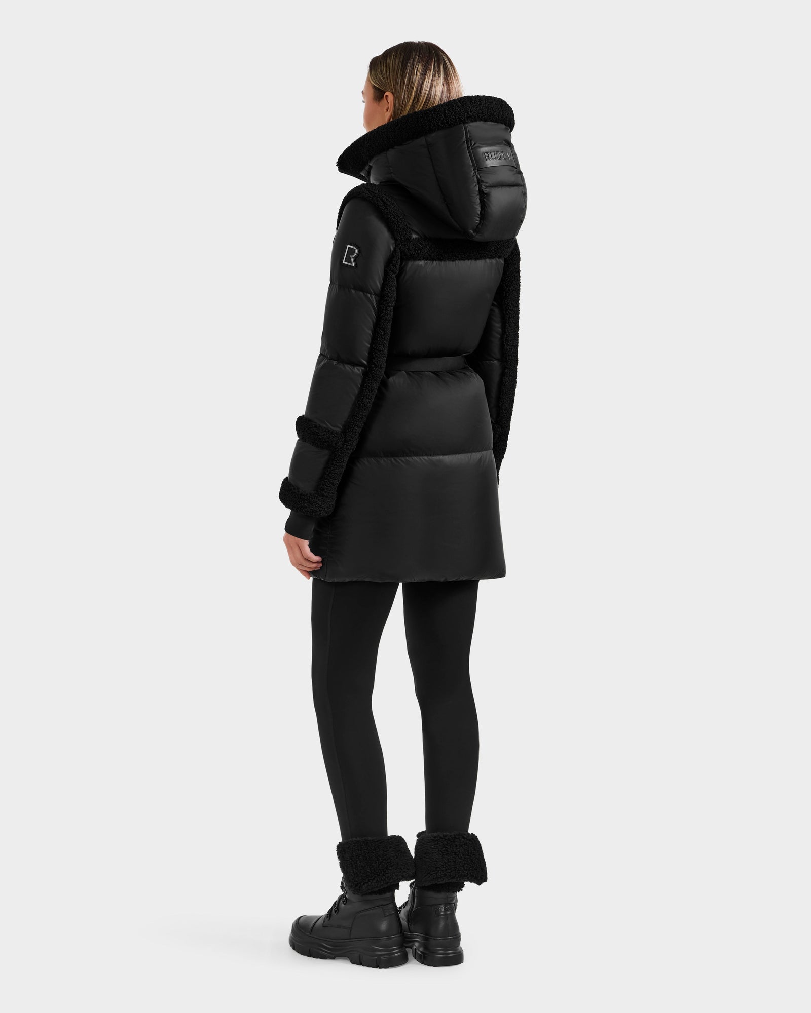 A person wearing the Rudsak JULIANA L - L125535 Black/Black down puffer with fur-lined hood, black leggings, and boots stands facing away against a light background—ideal for cold-weather performance.