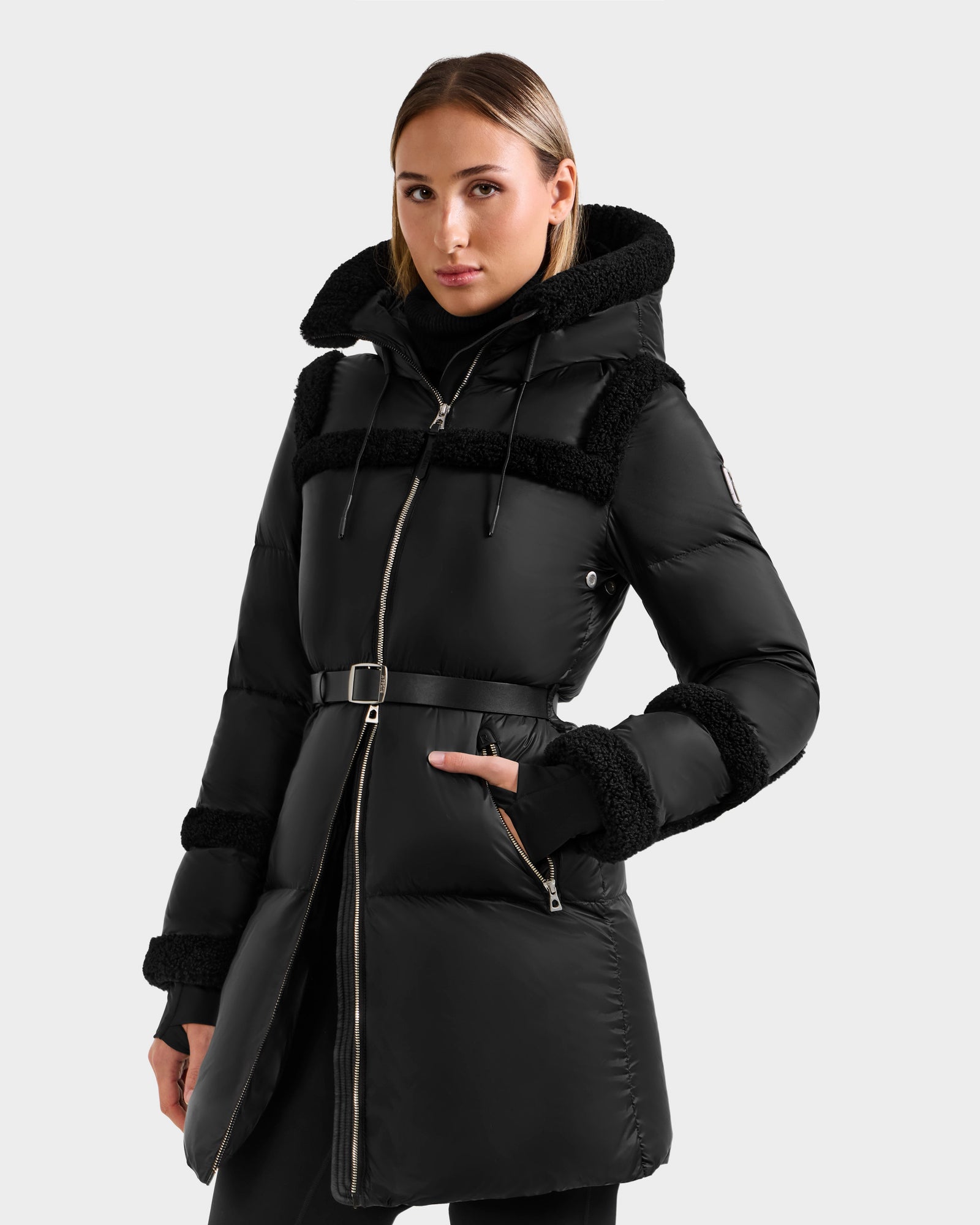 A woman wears the Rudsak JULIANA L - L125535 Black/Black puffer coat with faux fur trim and hood. She stands against a light gray background, posing with one hand in her pocket.