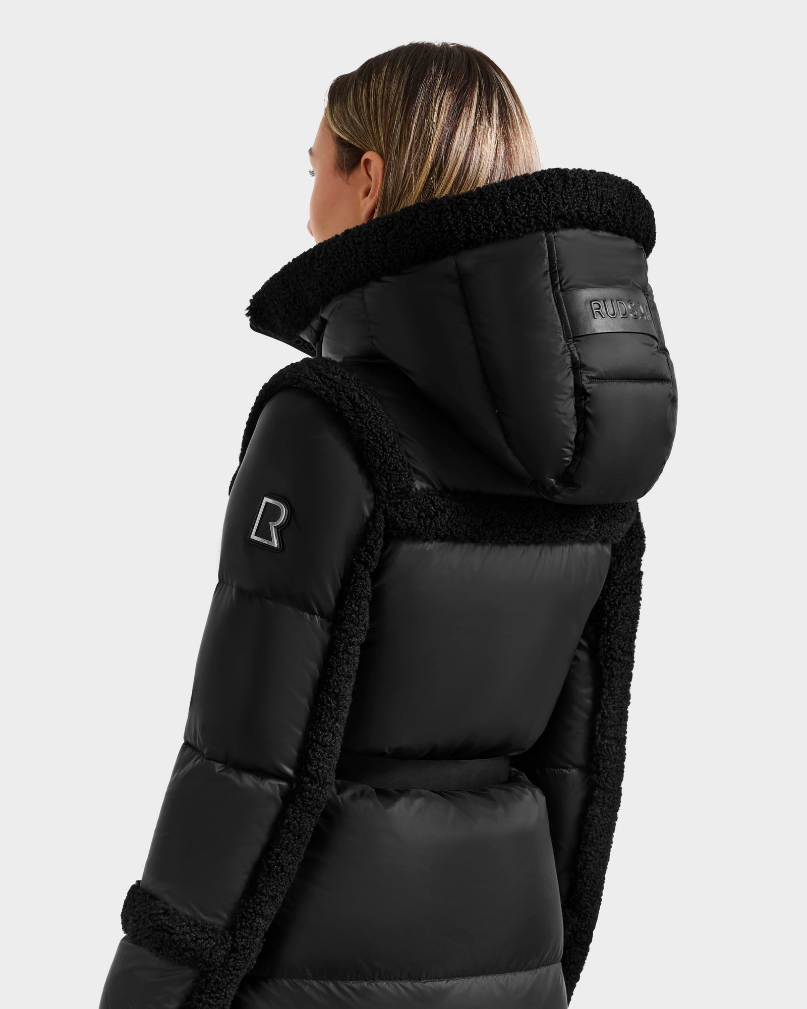 A woman wears the Rudsak JULIANA L - L125535 Black/Black quilted down puffer coat with a high collar, hood, soft trim, and logo patches on the sleeve and back—ideal for cold weather. She is shown from behind and slightly to the side.