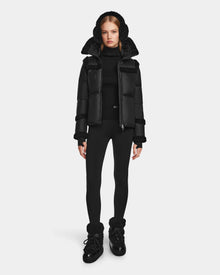 A woman faces forward, wearing the Rudsak JULIANA - L124509 Black/Black short down puffer jacket with a fur-lined hood, paired with a black turtleneck, leggings, and boots against a plain light background.