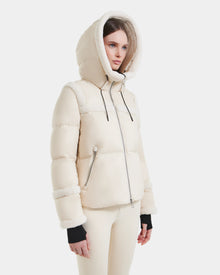 A woman wears the Rudsak JULIANA - L124509 Vanilla short down puffer jacket with a large hood and zippered pockets, paired with cream-colored pants against a plain white background.