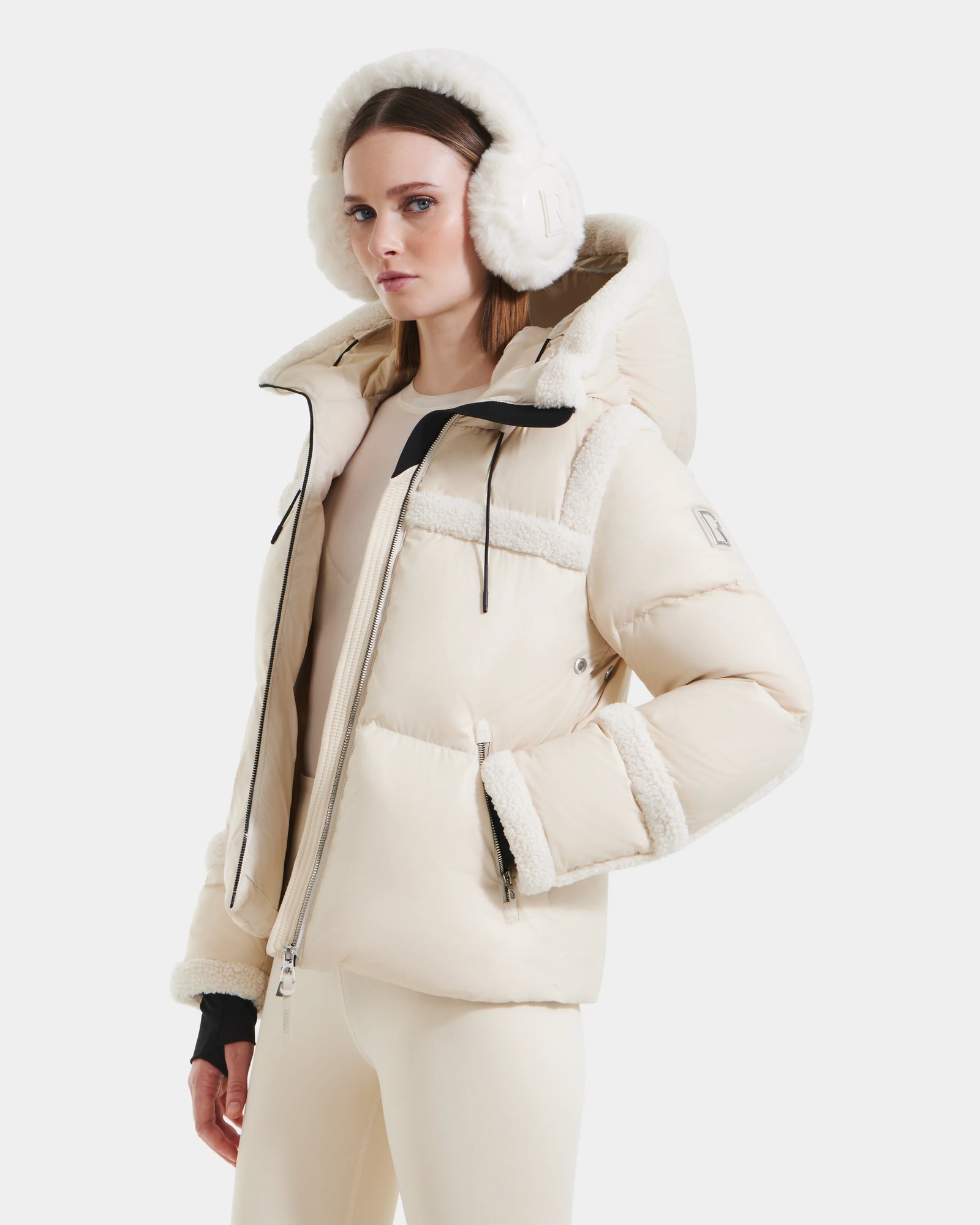 A woman models the Rudsak JULIANA - L124509 Vanilla short down puffer with faux shearling trim, paired with matching earmuffs and beige pants, standing confidently against a white background.