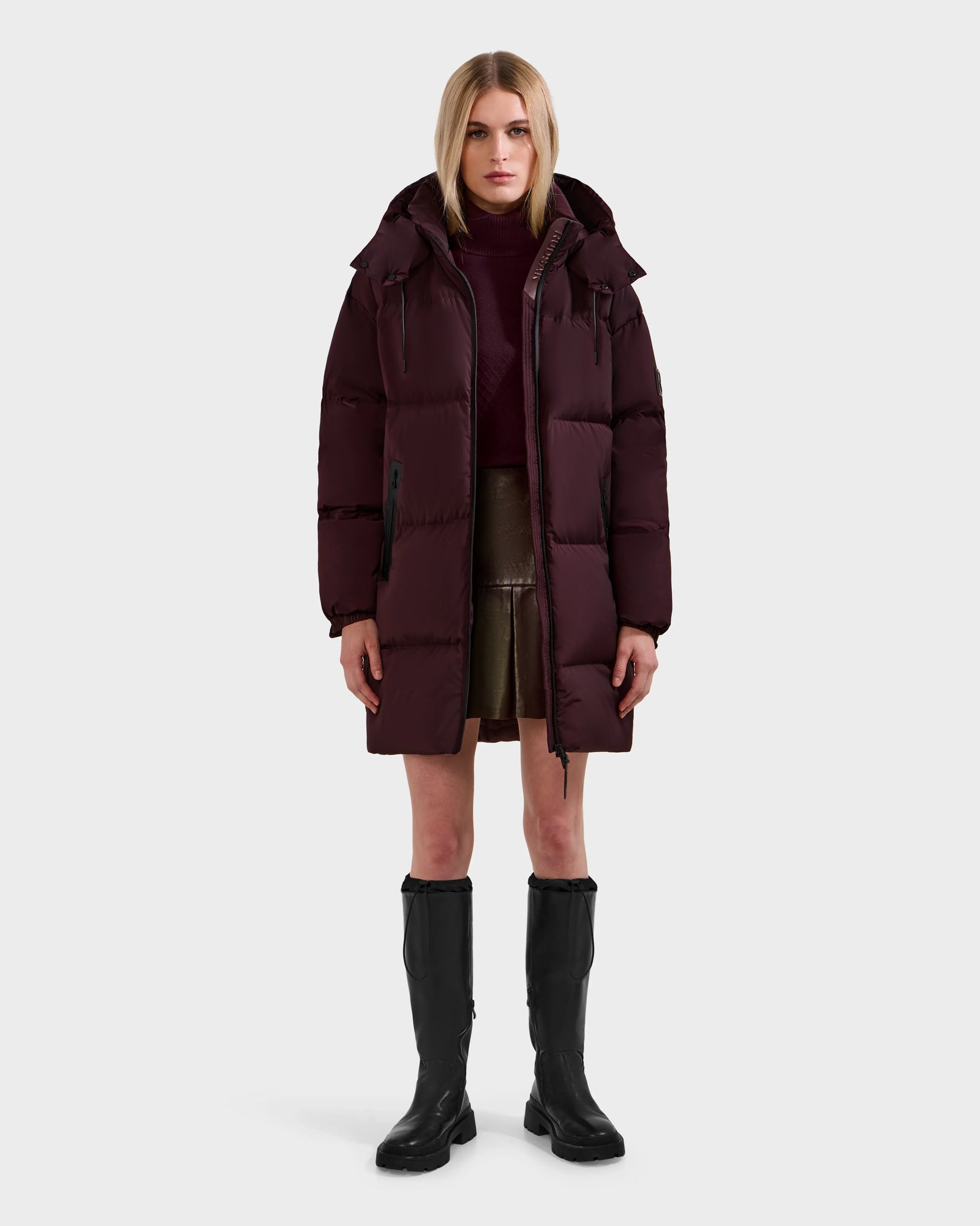 A woman faces forward in the Rudsak KARLEE L - 8125682 Bordeaux long down puffer, styled with a burgundy turtleneck, short brown skirt, and black knee-high boots—an easy cold-weather look set against a plain gray background.