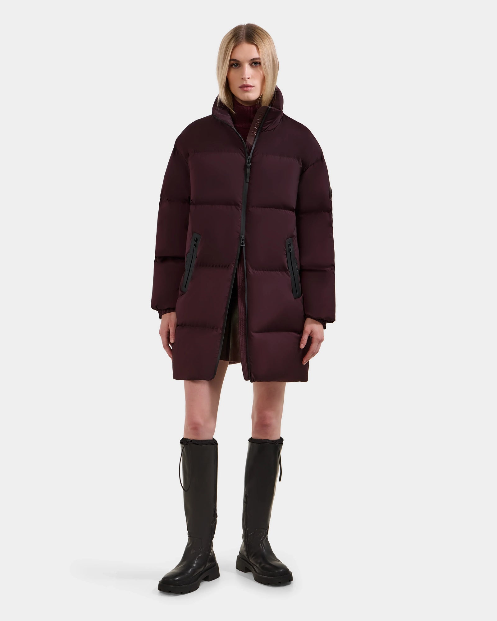 A woman stands against a white background wearing the Rudsak KARLEE L - 8125682 Bordeaux long-length down puffer jacket, paired with brown knee-high boots and a neutral expression—ideal for cold-weather wear.