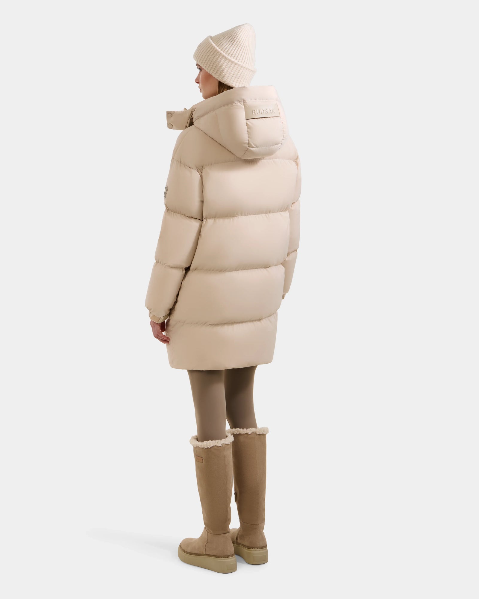 A person wearing the Rudsak KARLEE L - 8125682 Vanilla women’s long down puffer with vegan leather trims, tan leggings, beige boots, and a beige knit hat stands facing away against a white background. This jacket is stylish and cozy.