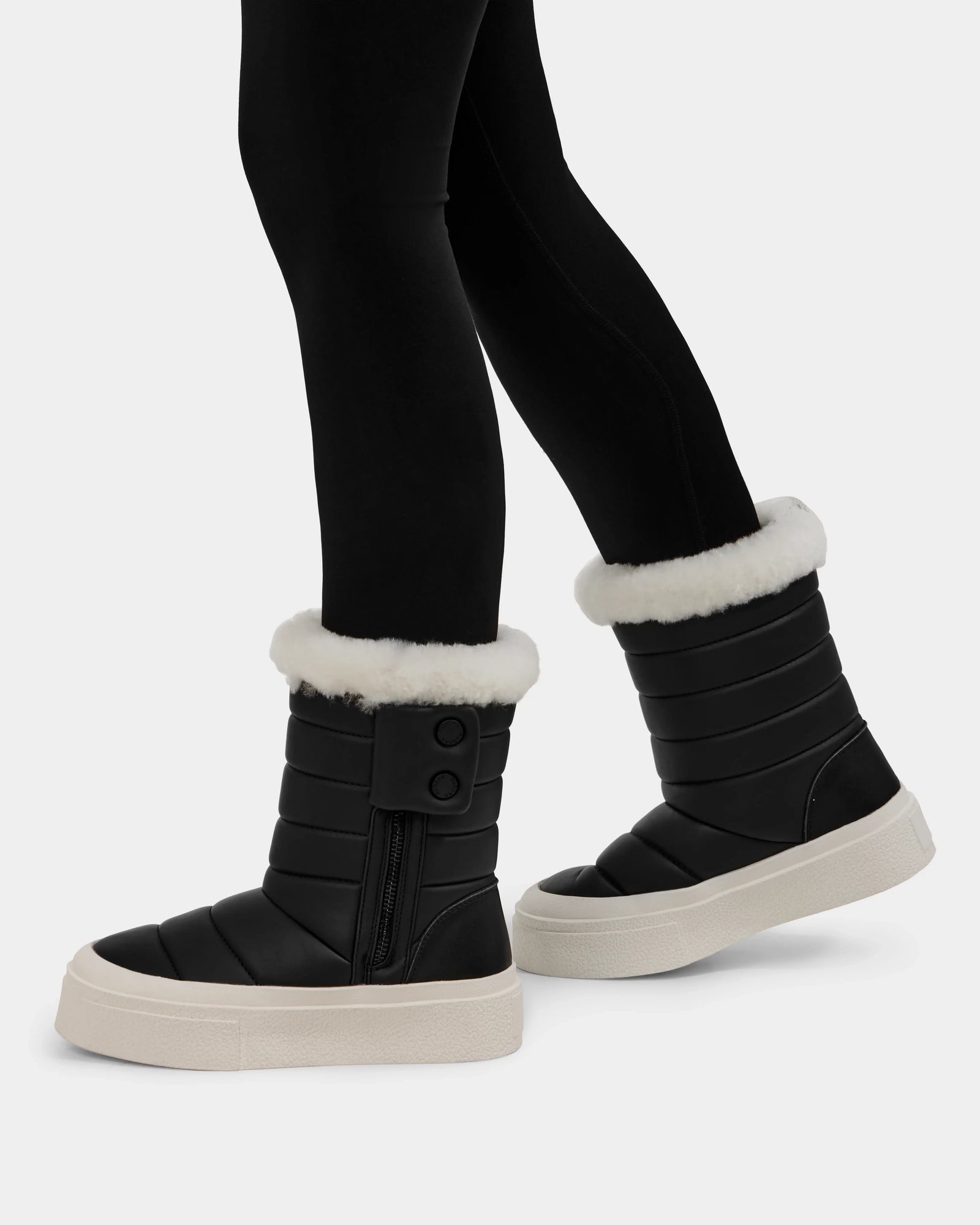 A person wears Rudsak KATHIE - L225516 Black quilted vegan leather mid boots with white faux fur trim, thick white soles, side zippers, and button details, paired with black leggings for a stylish winter look.