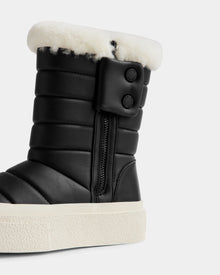 A close-up of Rudsak’s KATHIE - L225516 Black women’s mid boot, featuring black quilting, a side zipper, two buttons, white faux fur lining, and a thick white textured rubber sole.