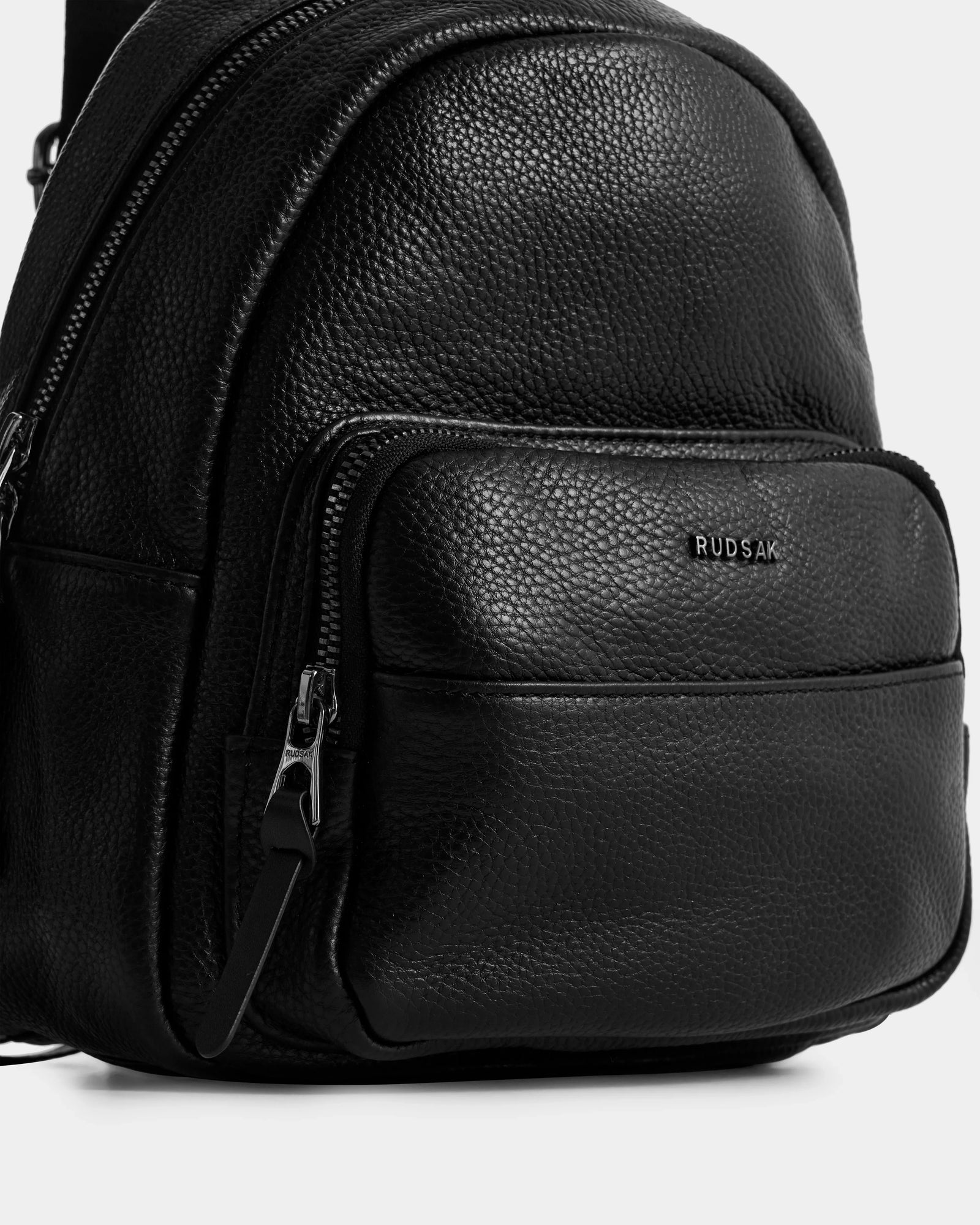 Close-up of the KATTY - L325014 mini backpack by Rudsak in black pebbled leather, with adjustable straps, a front zip pocket displaying the RUDSAK logo in silver, and visible zippers, shown against a white background.