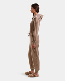 A woman stands in profile against a white background, wearing the Rudsak KETZY - L525503 Sand plush stretch velour hooded jumper with a drawstring waist and matching tan fuzzy slippers.