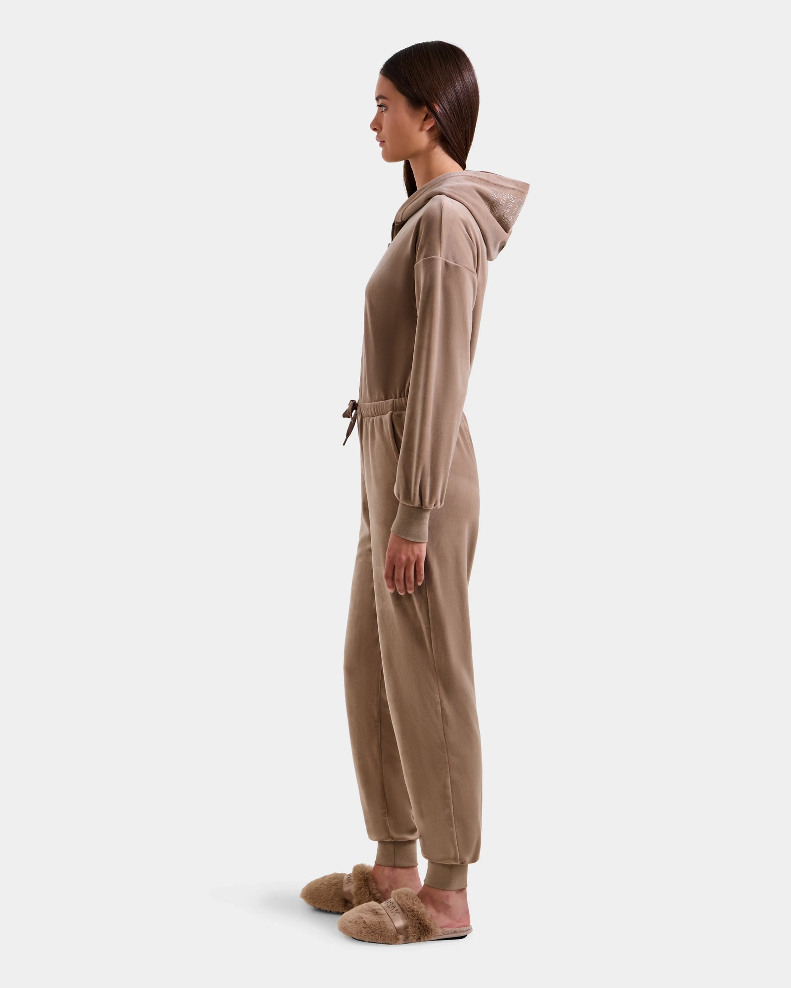 A woman stands in profile against a white background, wearing the Rudsak KETZY - L525503 Sand plush stretch velour hooded jumper with a drawstring waist and matching tan fuzzy slippers.