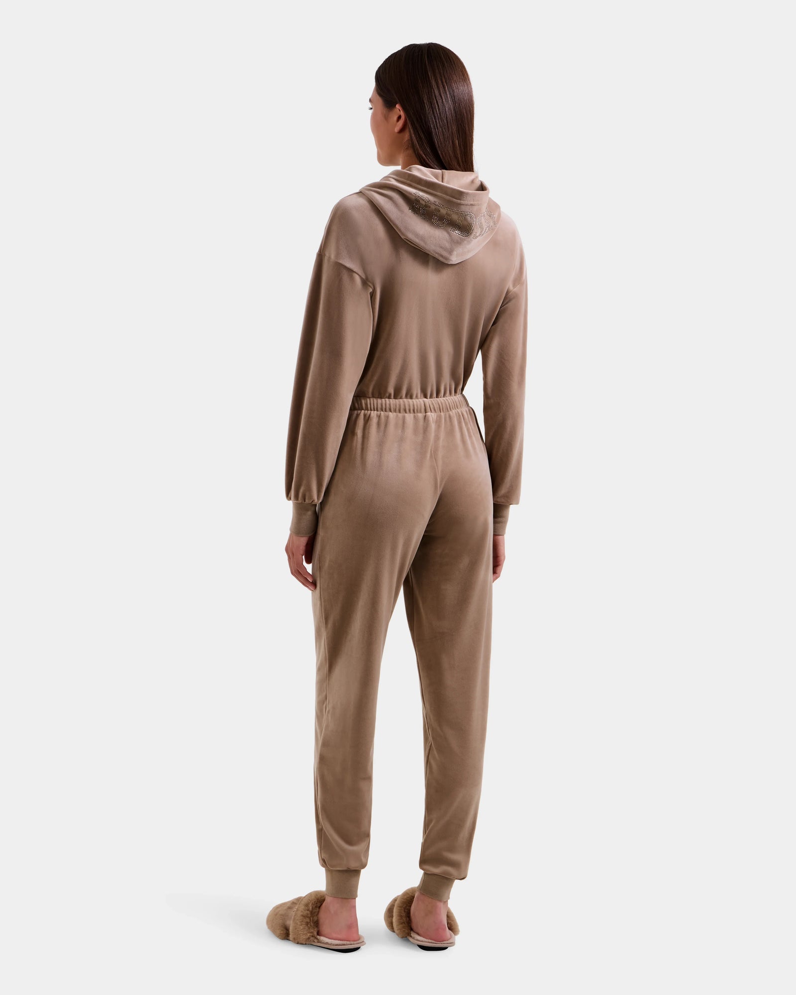A woman with straight brown hair stands facing away against a white background, wearing the Rudsak KETZY - L525503 Sand stretch velour hooded jumper with long sleeves and jogger cuffs, paired with fluffy slippers.