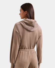 A woman with straight brown hair wears the Rudsak KETZY - L525503 Sand women’s velour hooded jumper, shown from the back, featuring crystal-sequin RUDSAK logo embellishments that add sparkle to its soft tan plush stretch velour.