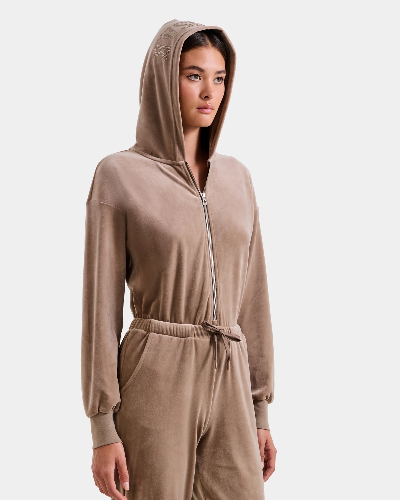 A woman wearing the Rudsak KETZY - L525503 Sand women’s velour hooded jumper, with plush stretch velour, an elastic waist, and a crystal-sequin Rudsak logo, stands against a plain light background.