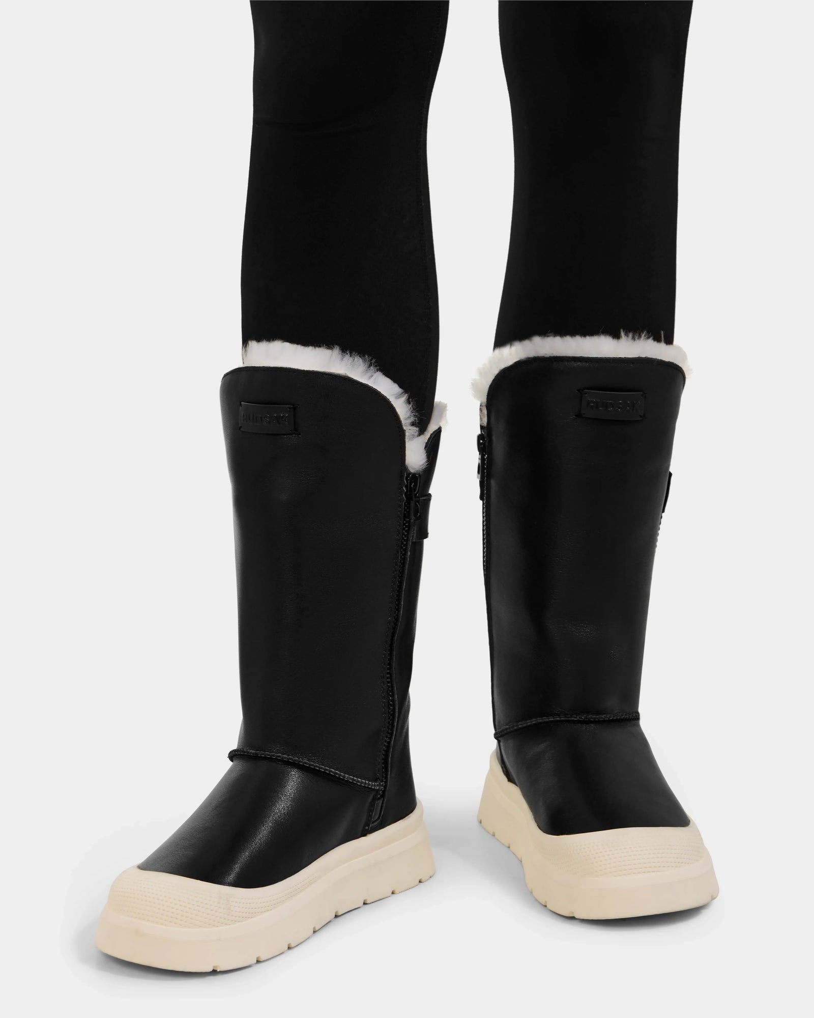 A person wears Rudsak’s KLAIR L - L225519 Black women’s leather boots with white fur lining, chunky white soles, side zippers, and a front label—styled with black leggings for perfect city walks.