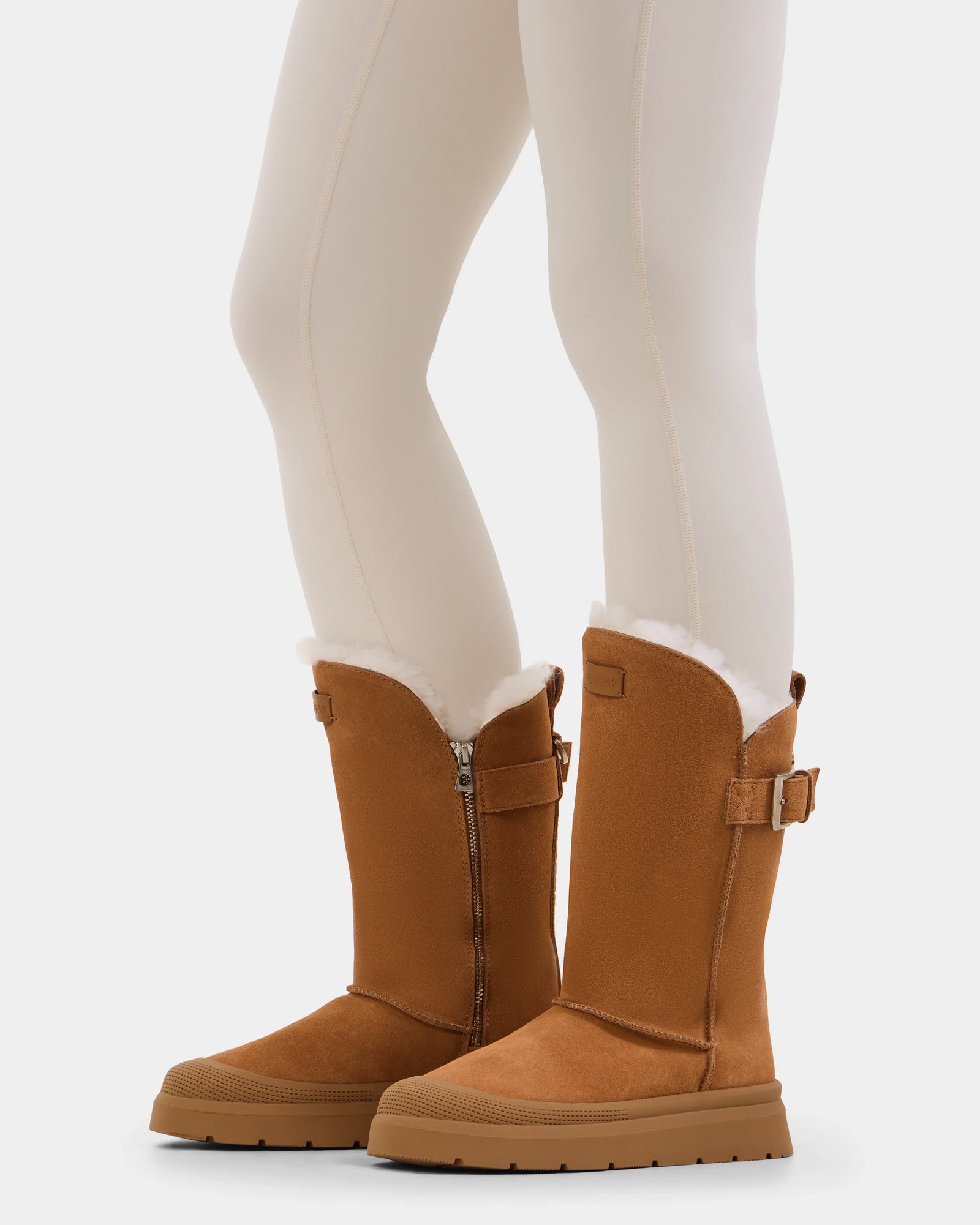 A person wearing white tights and Rudsak KLAIR - L225027 Tan women's suede winter boots with white fur lining, side zippers, and buckle straps; only their lower legs and feet appear against a white background.