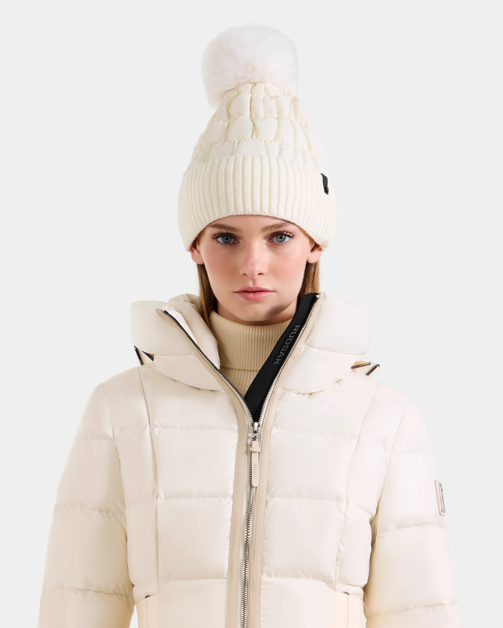 A woman faces forward against a plain light background, wearing the Rudsak LANYA - L423514 Iceland cream puffer jacket and a matching women's pom pom hat.
