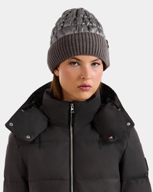 A woman faces forward with a neutral expression, wearing the Rudsak LANYA - L423514 Pewter knit beanie and a dark high-collar puffer jacket, set against a plain light gray background.