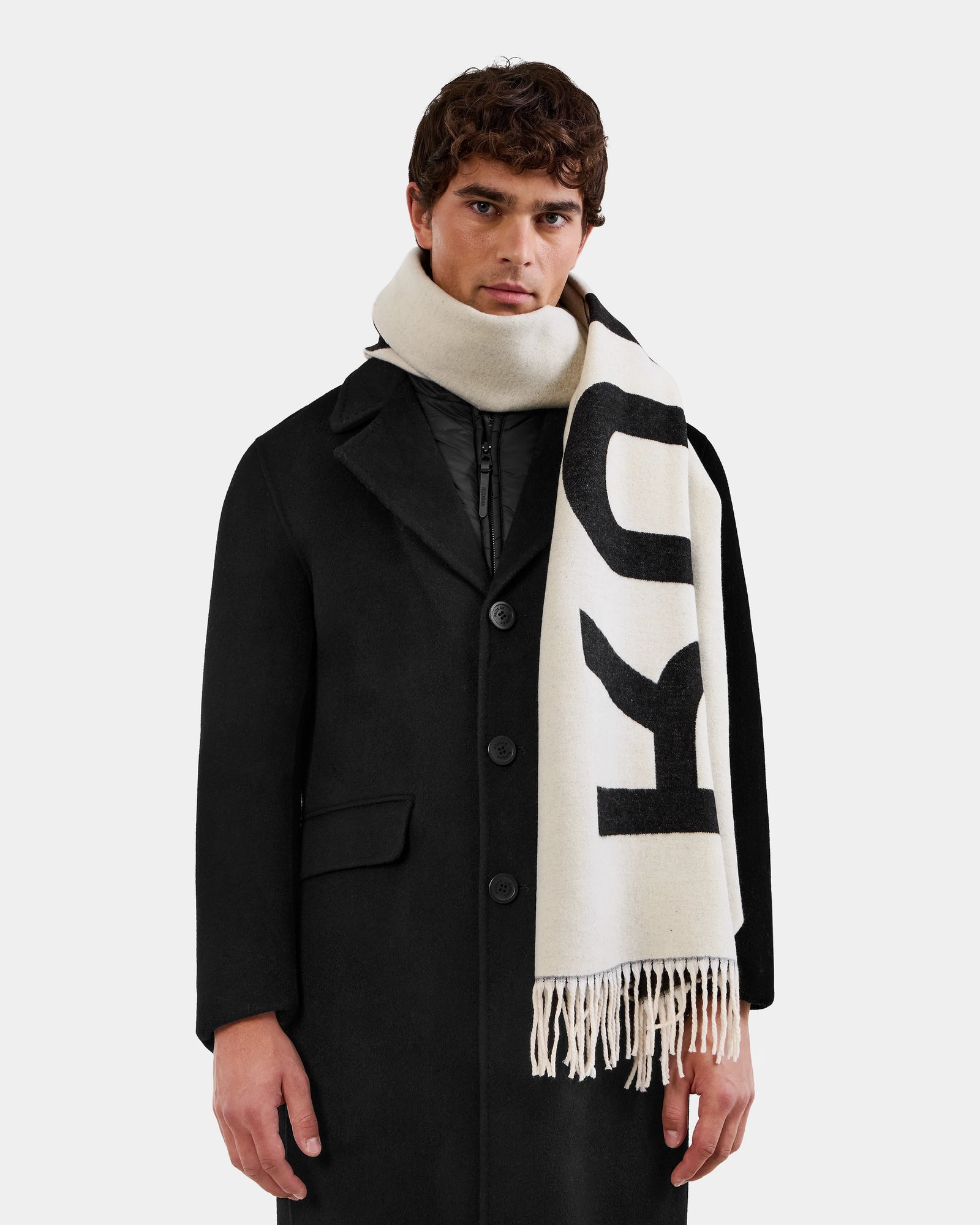 A person wearing the Rudsak LEV - U425506 Black coat and a cream-colored scarf with large black RUDSAK lettering and fringed ends, wrapped around their neck, stands against a plain white background.