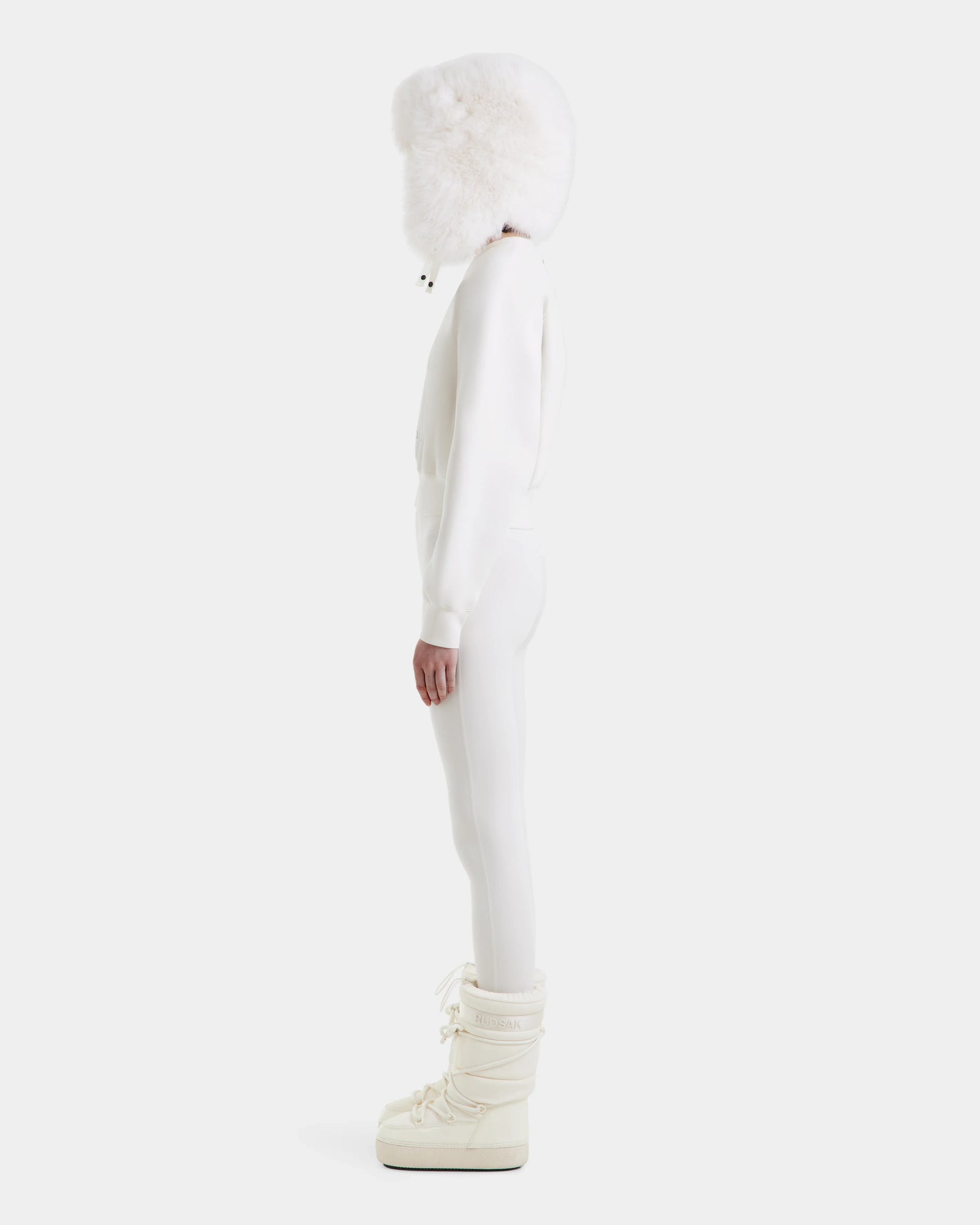 A person stands in profile wearing Rudsak's LIDDIE - L525505 Vanilla, a fitted all-white winter outfit with a fluffy white hood and tall off-white boots, set against a plain light gray background.