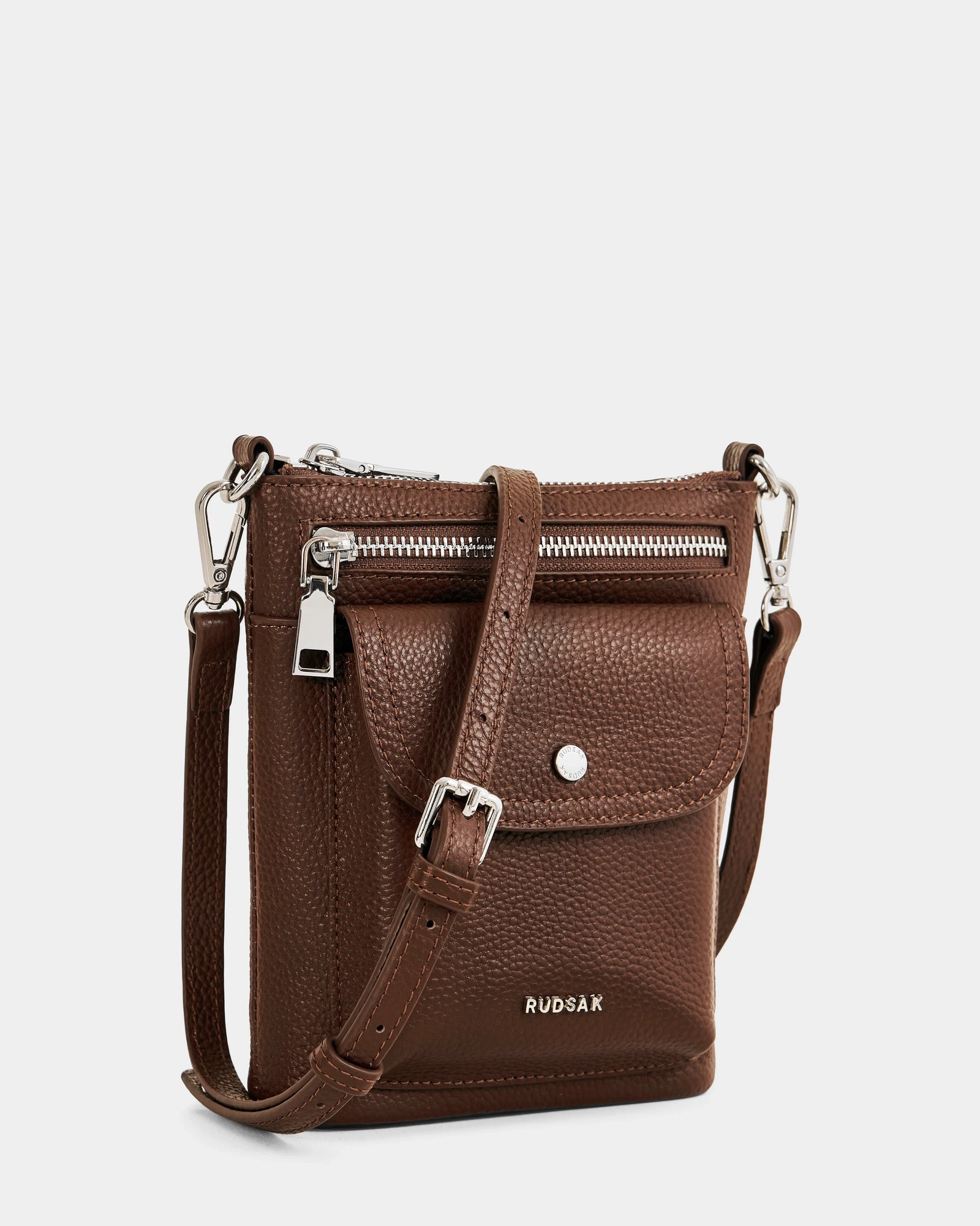 Rudsak LILA - L325513 Mocha is a small brown women's crossbody bag made of pebbled leather, featuring a front zip pocket, flap pocket with snap button, adjustable strap, silver hardware, and the Rudsak logo in silver near the bottom.