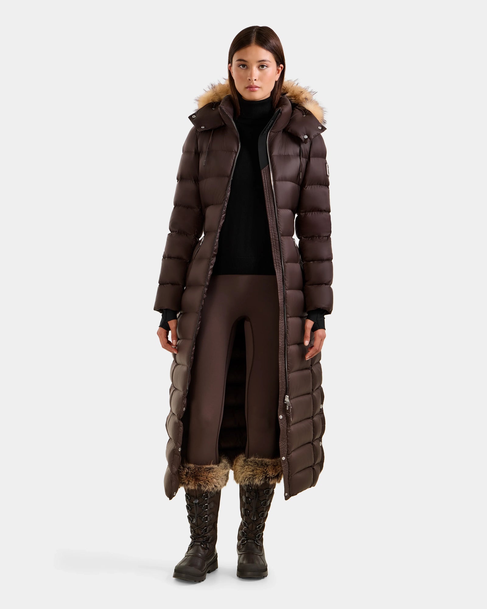 A woman wears the Rudsak LILITH L - L125528 Espresso maxi down puffer with RDS-certified filling and a fur-trimmed hood, paired with a black turtleneck, brown side-slit pants, and tall fur-detailed lace-up boots against a plain white backdrop.