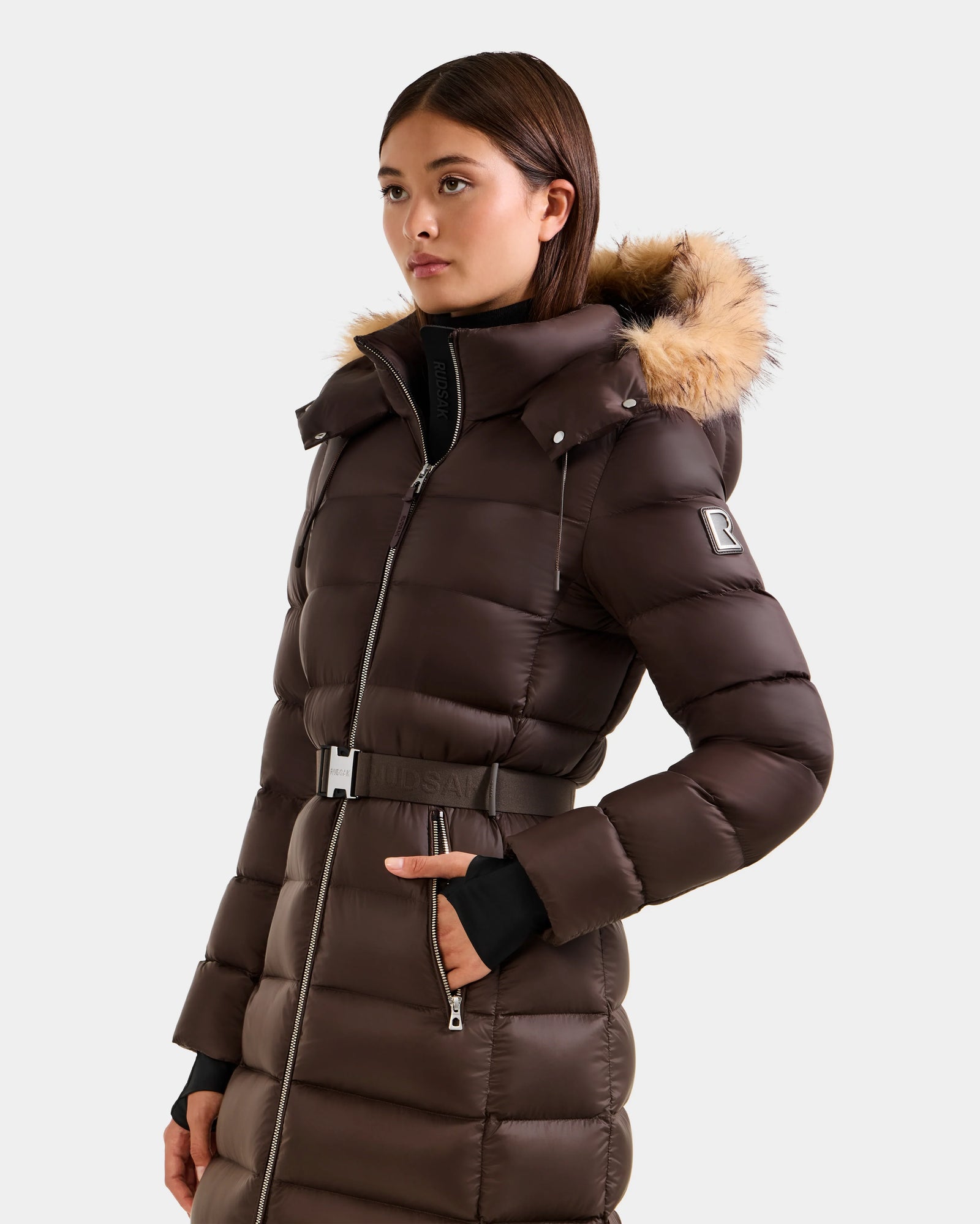 A woman wears the Rudsak LILITH L - L125528 Espresso maxi down puffer coat with a faux-fur trim hood, belted waist, zipper, side pockets, and sleeve logo patch, standing against a plain light background.