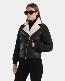 A woman wearing black sunglasses and black pants models the Rudsak MALA - 8125628 Black women’s shearling jacket, featuring a white collar and cropped silhouette, standing against a plain white background.