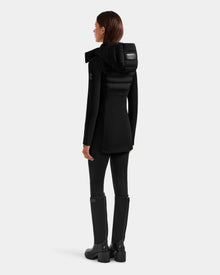 A woman faces away, wearing the sleek black Rudsak MARRY M - L125519 women’s mid-length hybrid coat with black pants and knee-high boots against a plain light gray background.