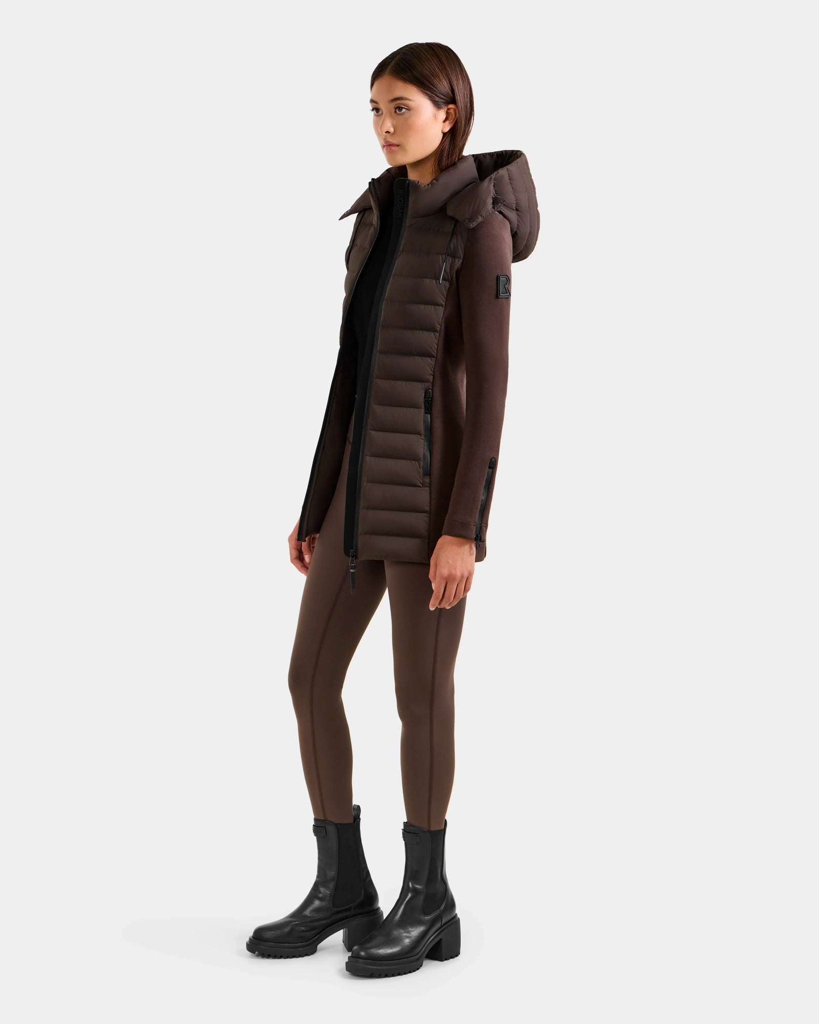 A woman faces slightly left, wearing Rudsak's MARRY M - L125519 Espresso long fitted brown puffer coat with down insulation and removable hood, brown leggings, and black ankle boots against a plain light background.