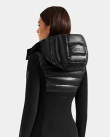 A woman with straight brown hair is seen from behind, wearing the Rudsak MARRY S - L125521 Black jacket with a shiny, padded hood and back panel. Weather-resistant details and the Rudsak logo are on the hood and back. The background is plain white.