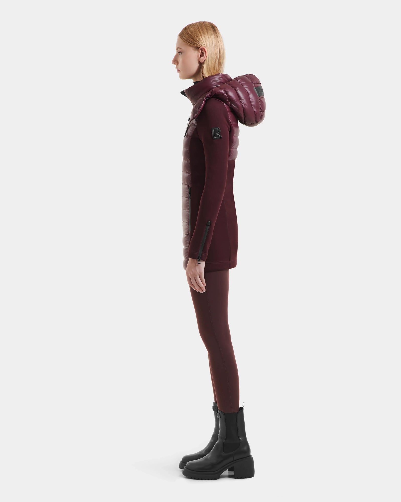 A person with straight blonde hair stands in profile, wearing the Rudsak MARRY S - L125521 Wine weather-resistant hooded puffer vest, burgundy leggings, and black chunky boots against a plain white background.