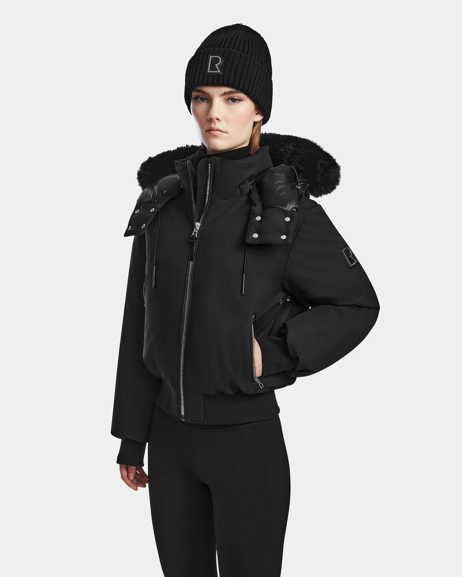 A woman wears the Rudsak MAUDE - L124522 Black/Black down bomber with silver buttons and a fur-lined hood, plus a black Rudsak beanie, standing against a plain white background.