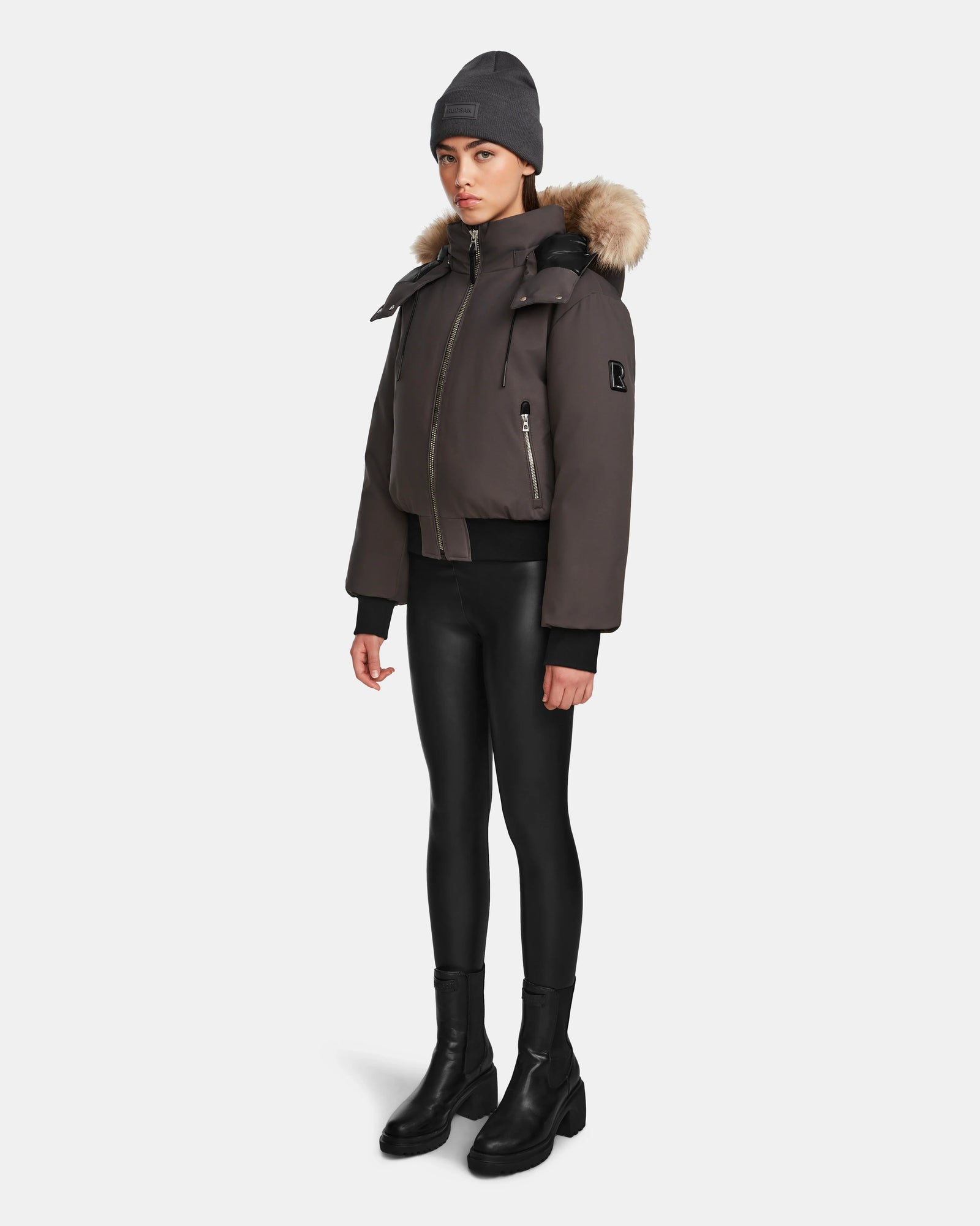 A person models the Rudsak MAUDE - L124522 Grey beanie, pairing it with a classic winter jacket with a fur-lined hood, black shiny leggings, and black boots against a plain white background.