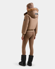 A person wearing the Rudsak MAUDE - L124522 Sand women's down bomber jacket with matching tan leggings, a knit beanie, and black winter boots stands facing away from the camera against a plain white background.