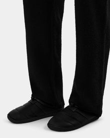 A person wearing black textured pants and Rudsak MILTON - M225504 Black minimalist men’s quilted slippers with faux-shearling lining on a plain white background.