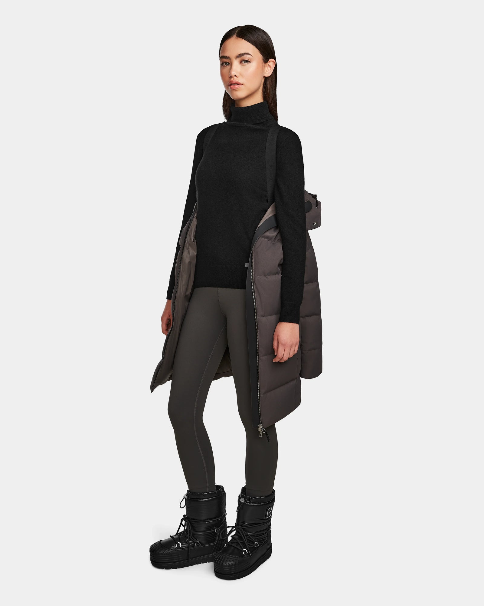 A woman stands against a plain background wearing Rudsak’s MIRIAM - L123544 Grey women’s down puffer coat with a weather-resistant shell, paired with a black turtleneck sweater, dark gray leggings, and bulky black winter boots.