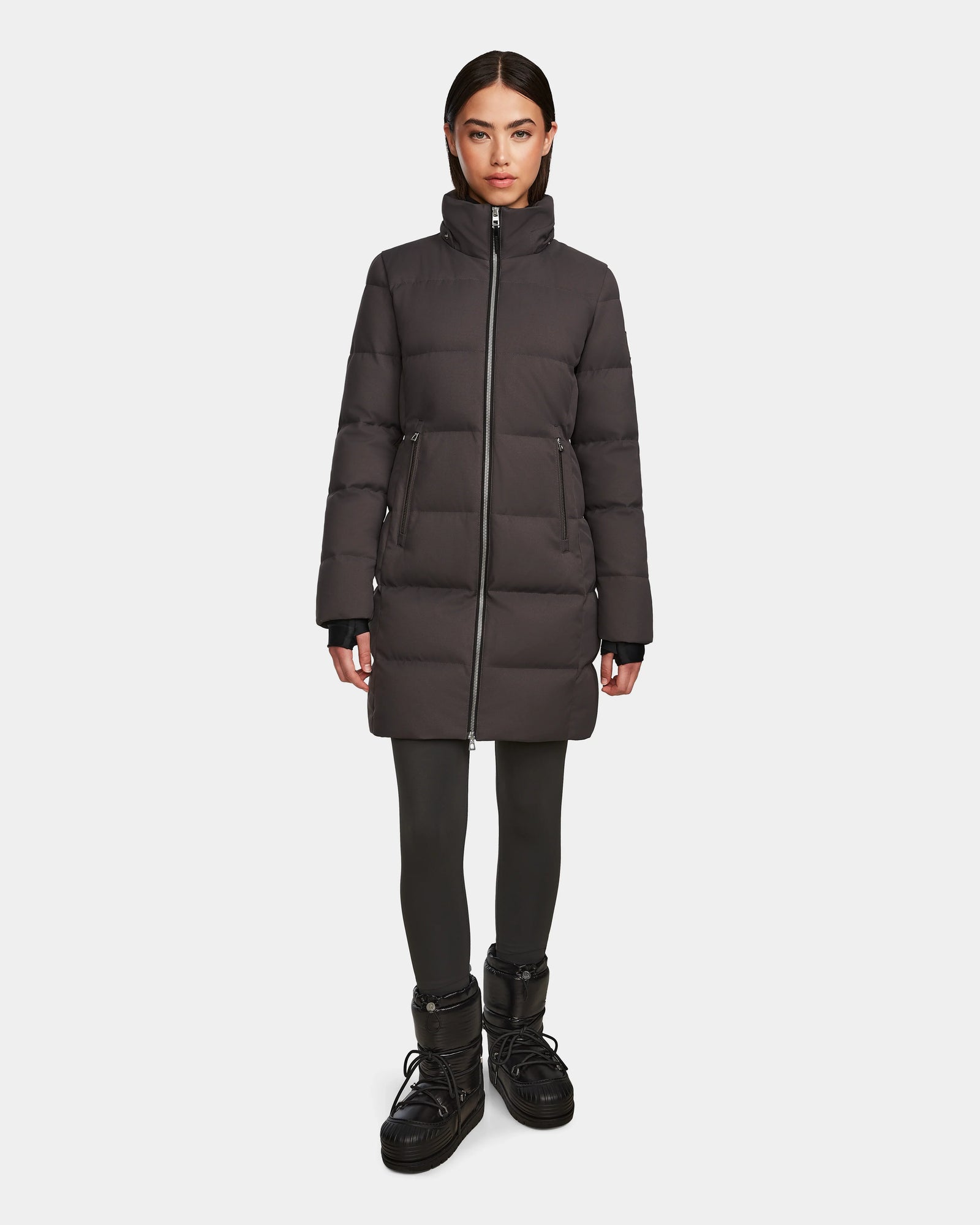 A woman faces forward, wearing the Rudsak MIRIAM - L123544 Grey women’s down puffer jacket with a high collar, paired with black leggings and winter boots against a plain white background—an essential winter piece.