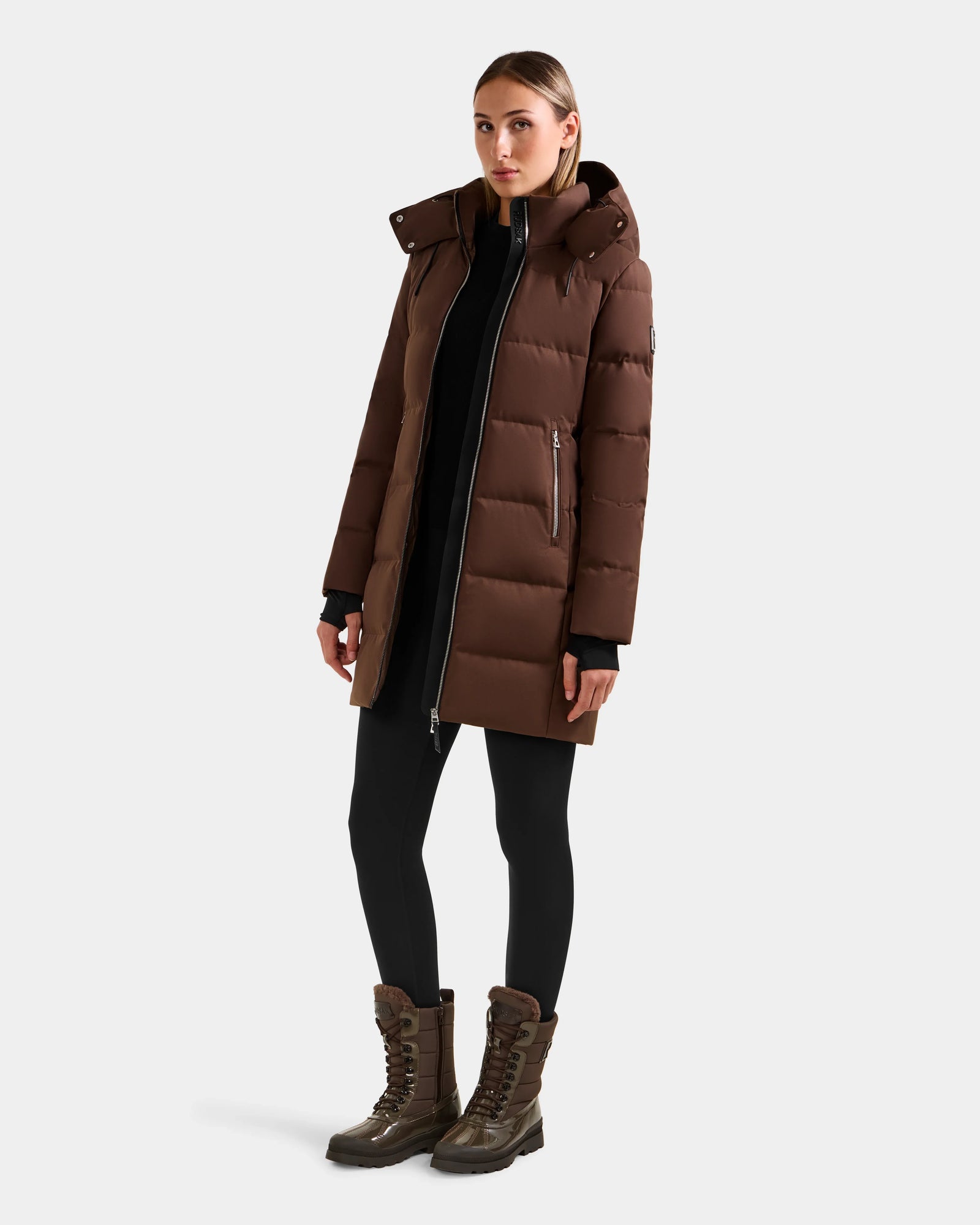 A woman faces forward wearing the Rudsak MIRIAM - L123544 Mocha, a women’s down puffer coat with a weather-resistant shell, paired with black leggings and brown lace-up boots against a plain light background.