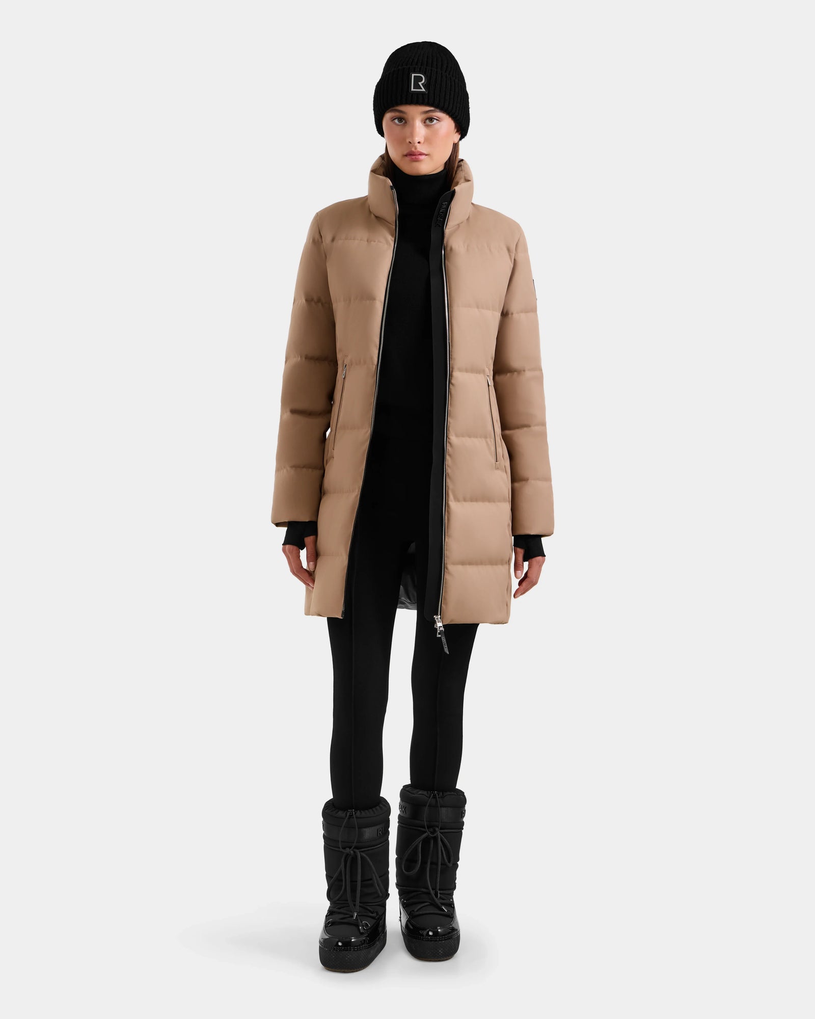A person stands facing forward, wearing the Rudsak MIRIAM - L123544 Sand weather resistant women’s down puffer, black leggings, black winter boots, gloves, and a logo beanie against a plain light gray background.
