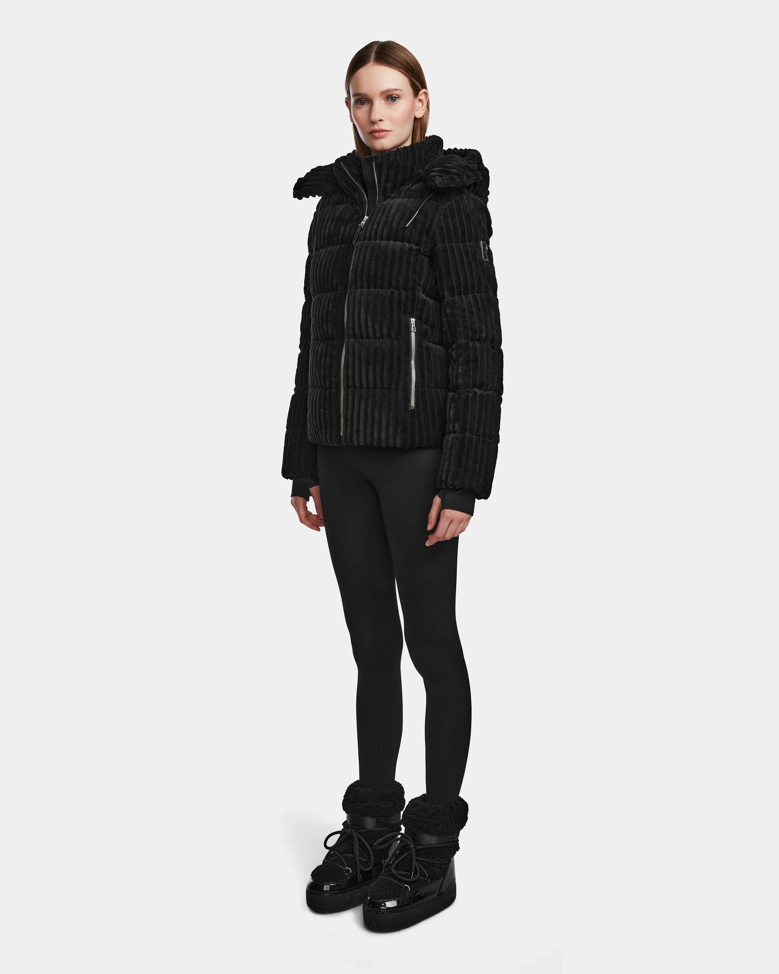 A woman with straight brown hair and a neutral expression stands against a plain background wearing the MIRRY FF - L124507 Black womens short down puffer jacket, paired with black leggings and winter boots.