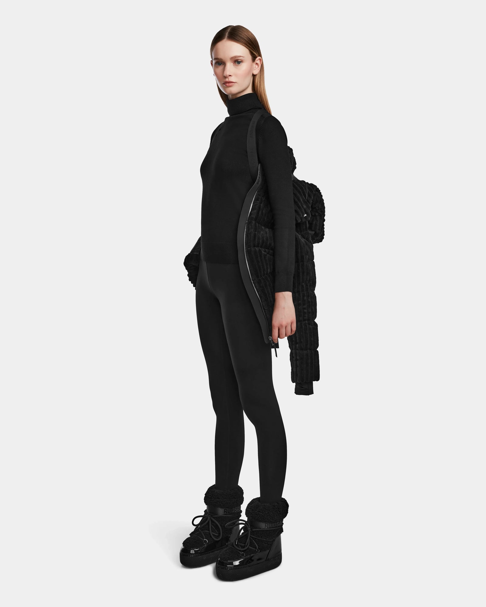 A woman with straight brown hair stands in profile, holding the MIRRY FF - L124507 Black womens short down puffer jacket over her left arm, paired with a black turtleneck, leggings, and boots against a plain white background.