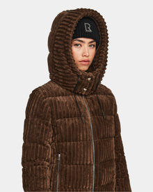 A woman wears the Rudsak MIRRY FF - L124507 Espresso short down puffer jacket with a hood and black beanie. The brown, corduroy-style quilted coat features a zippered front. She stands slightly turned against a plain background.