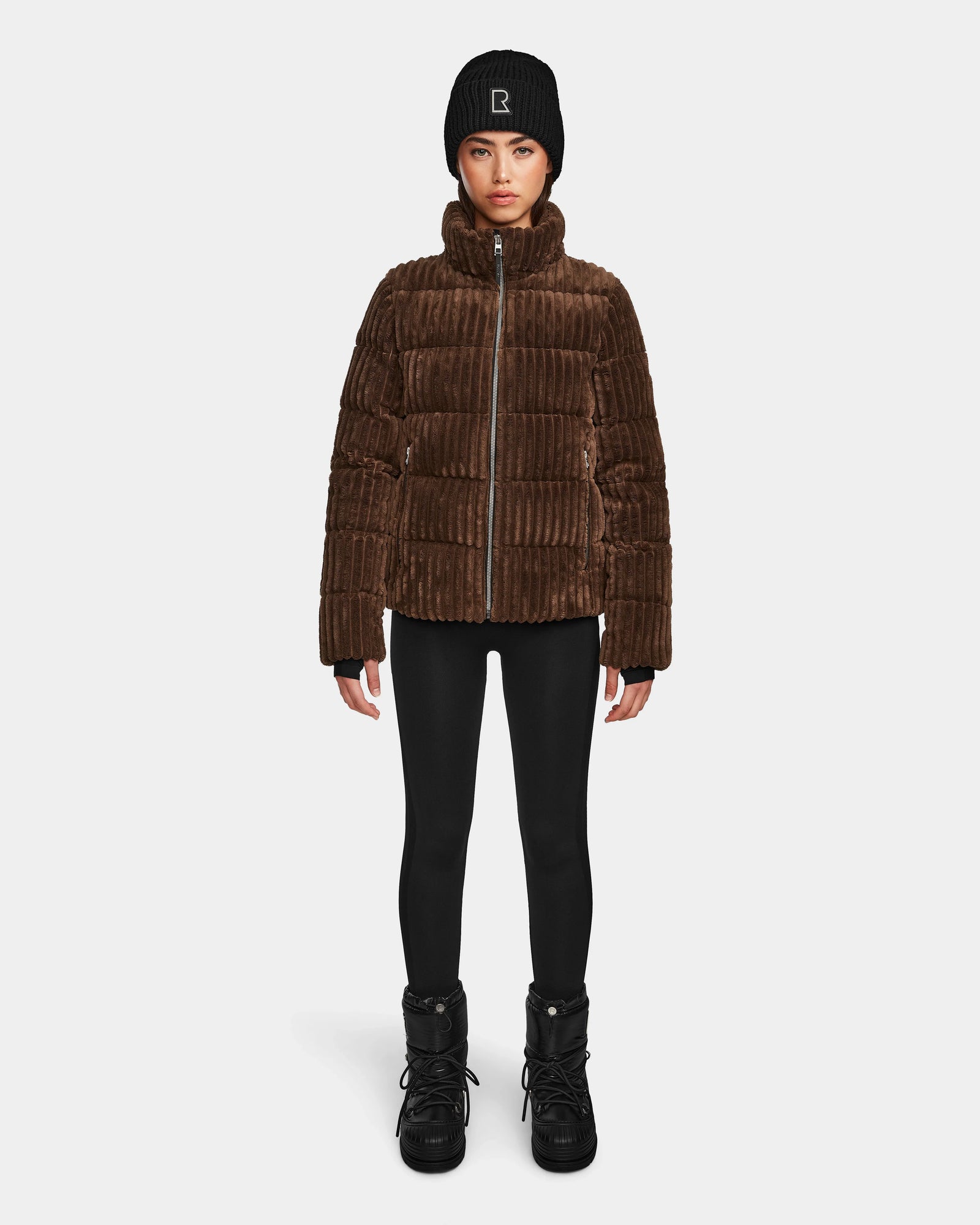 A person wearing the Rudsak MIRRY FF - L124507 Espresso women's short down puffer jacket, black leggings, black winter boots, and a black beanie stands facing forward against a plain white background.