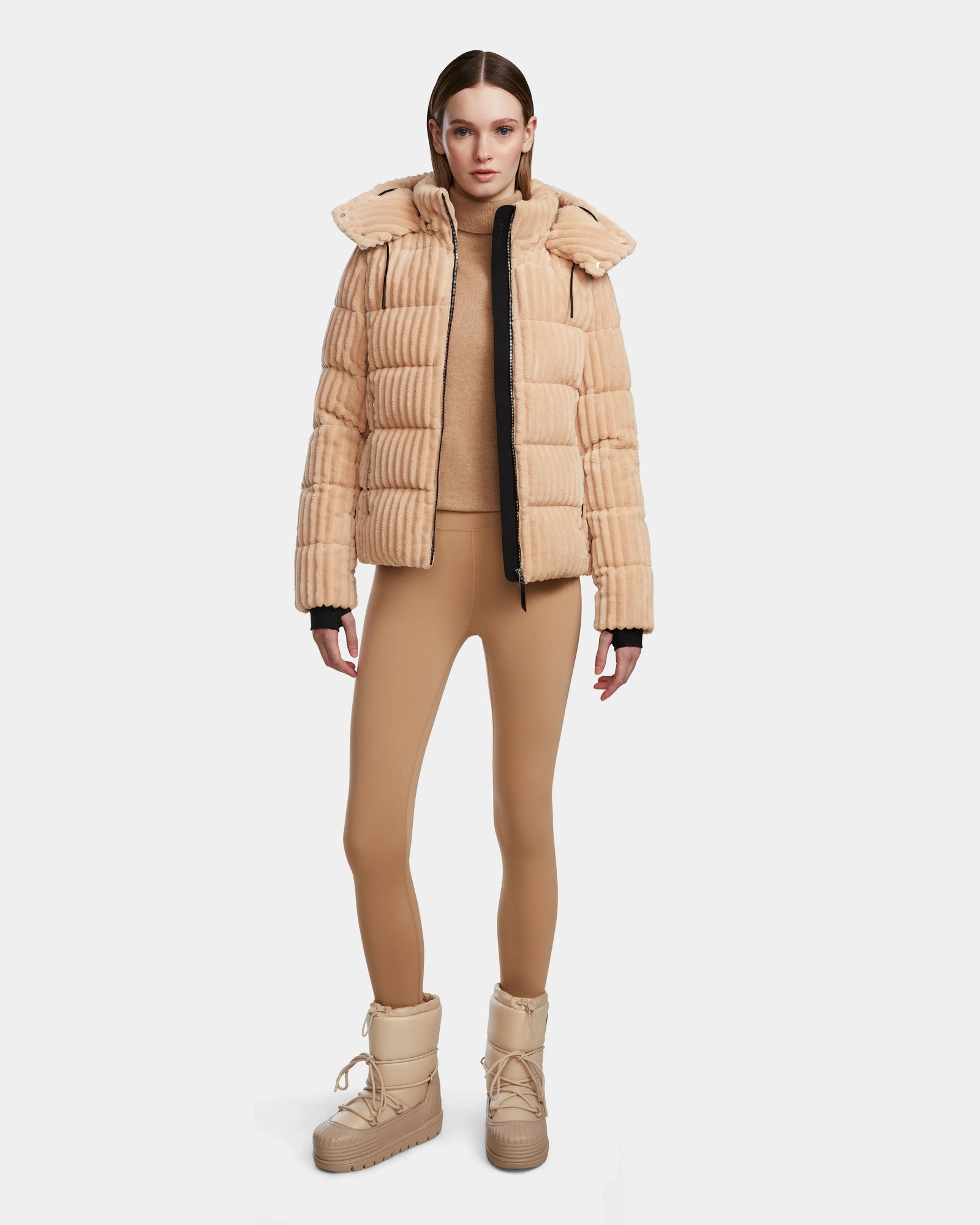 A woman faces forward wearing the Rudsak MIRRY FF - L124507 Iceland beige women’s short down puffer jacket, a tan turtleneck, tan leggings, and beige winter boots against a plain white background.