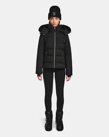A person stands facing forward against a plain white background, wearing the Rudsak MIRRY M - L124533 Black women’s quilted down coat with fur-trimmed hood.